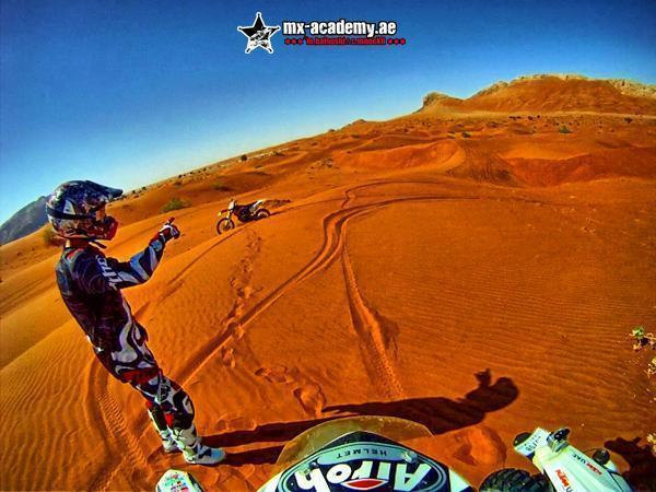 Desert bike tour Dubai: seen from the handlebars, a rider in the sand raises an arm while another bike waits further up