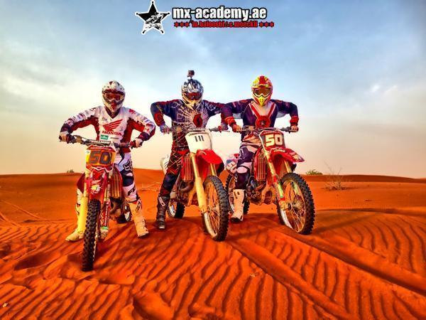 Desert motorbike tour Dubai: three riders sit on red Honda bikes side by side on rippled red sand under clouds