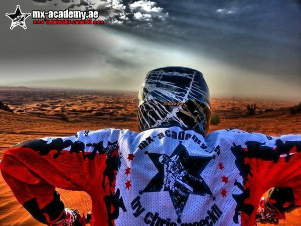 Dirt bike desert tour Dubai: a rider in an MX-Academy jersey looks out over wide dunes under a cloudy sky
