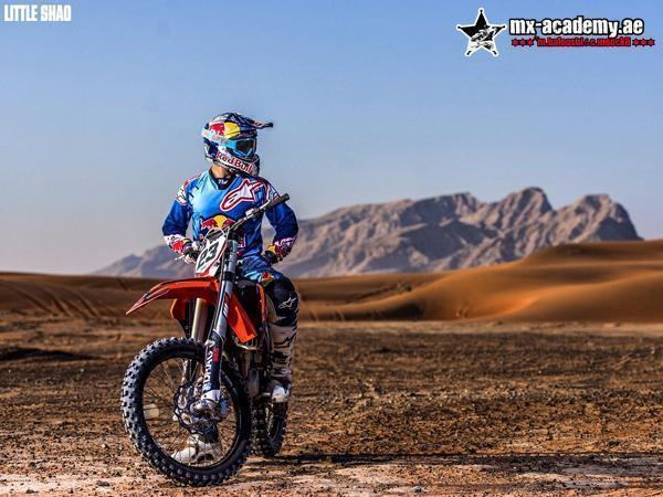 Dirt bike UAE: a rider in blue Red Bull kit waits on an orange bike, dunes and a rocky mountain behind