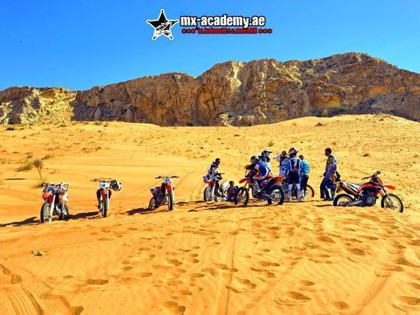 Enduro desert tour Dubai: riders gather around their parked bikes on pale sand below a sunlit rocky cliff