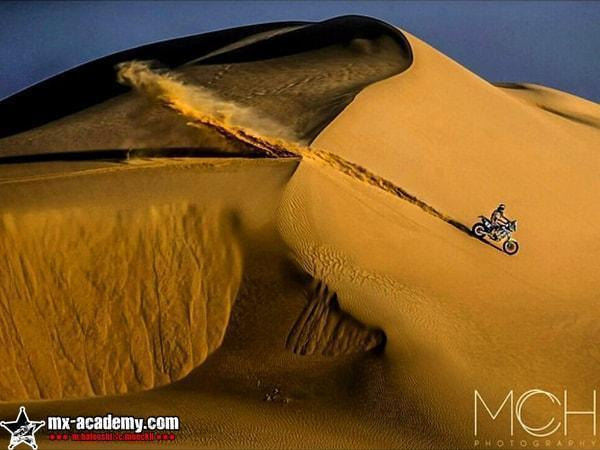 Enduro Dubai dunes: a lone rider crosses the face of a huge golden dune, sand blowing off its sharp crest