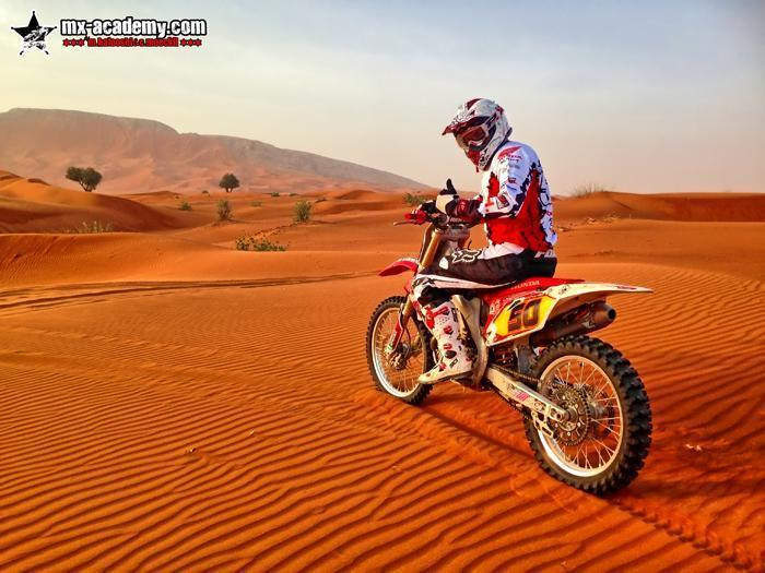 Enduro Dubai: a rider in red and white Honda kit sits on his bike on rippled red sand, rocky hills behind