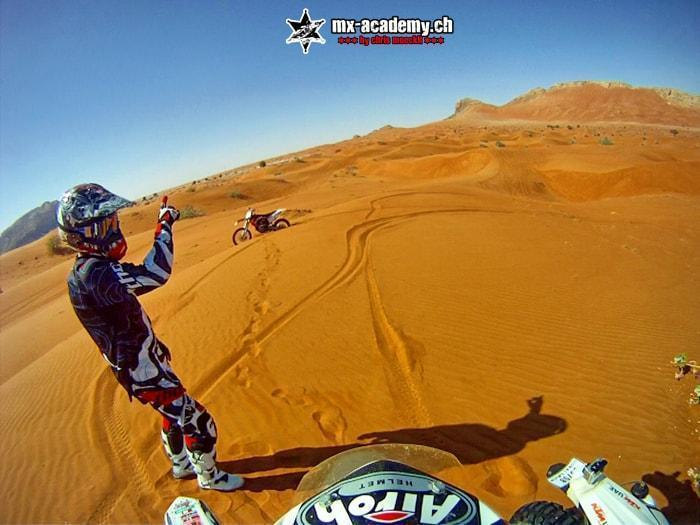 Enduro in Dubai: a rider on a dune raises his arm, a second bike parked in the tracks, seen from the handlebars