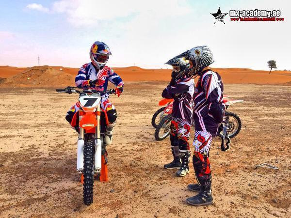 Kids dirt bike Dubai: rider on an orange KTM talks to two young riders in helmets, red dunes in the background