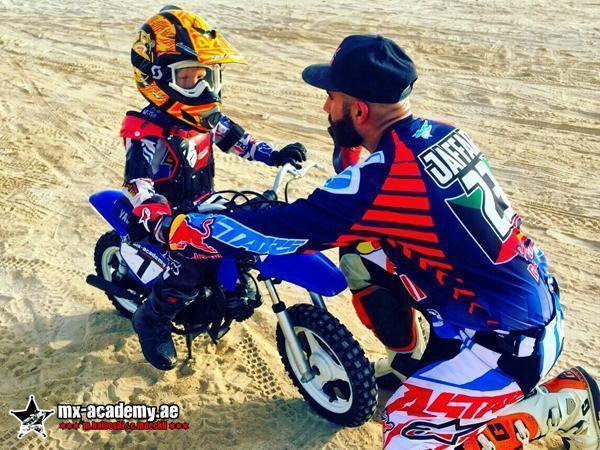 Kids motocross Dubai: coach in a cap kneels next to a small child in an orange helmet on a blue kids' bike
