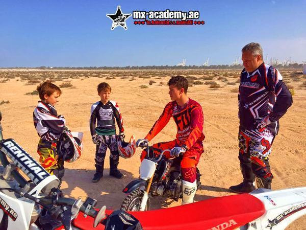 Kids motocross Dubai: two boys in riding gear hold their helmets next to a young rider on a small bike and an adult