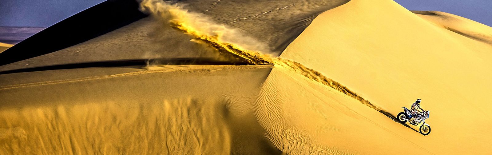 Motocross Dubai: a rider crosses huge golden sand dunes on an enduro bike, a trail of sand blowing off the crest