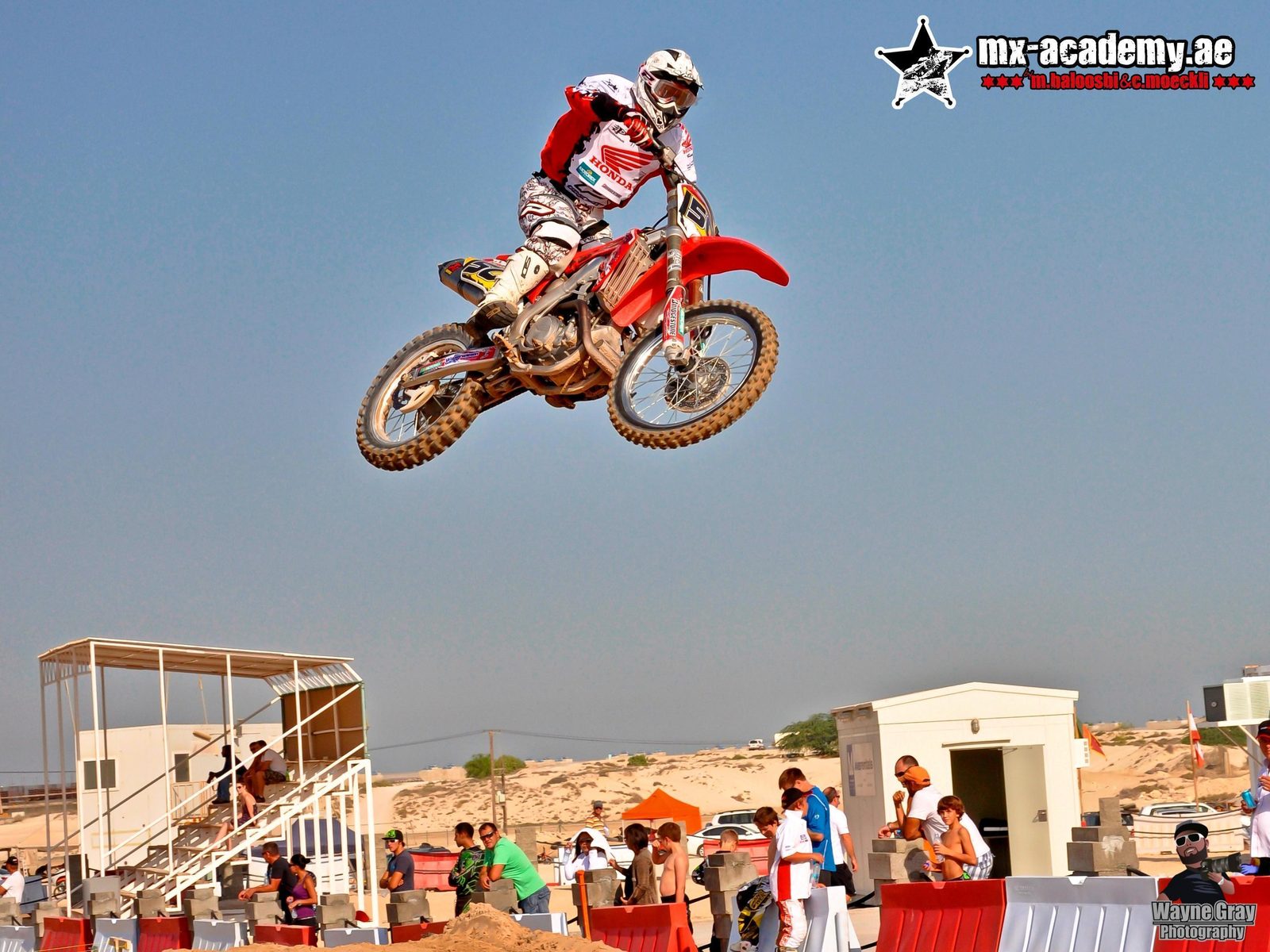 Motocross Dubai: rider in a red and white Honda jersey jumps high above the track, spectators and a stand below