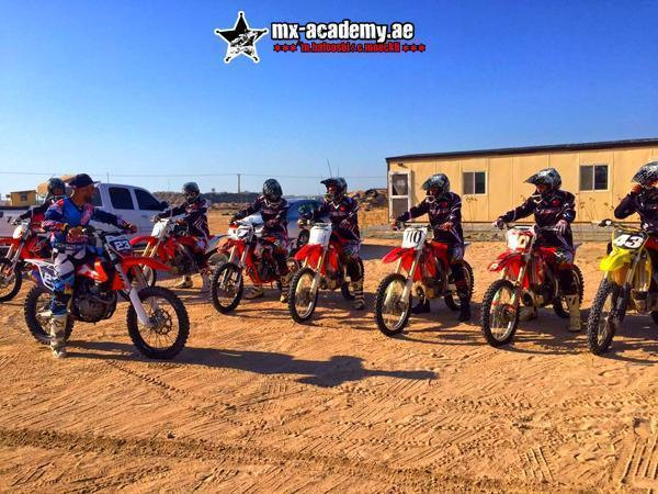 Motocross in Dubai: a row of riders in helmets waits on their bikes on sandy ground next to a low building