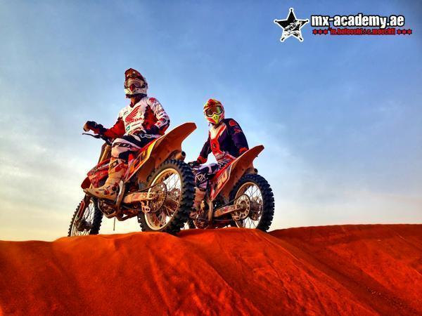 MX Dubai: two riders sit on orange motocross bikes on the crest of a red sand dune under a clear sky